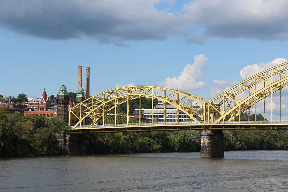 Photo by Frankie Bonamo, 2021. Features one of the many yellow bridges of Pittsburgh with some of the smokestacks left from the city's industrial period on the left