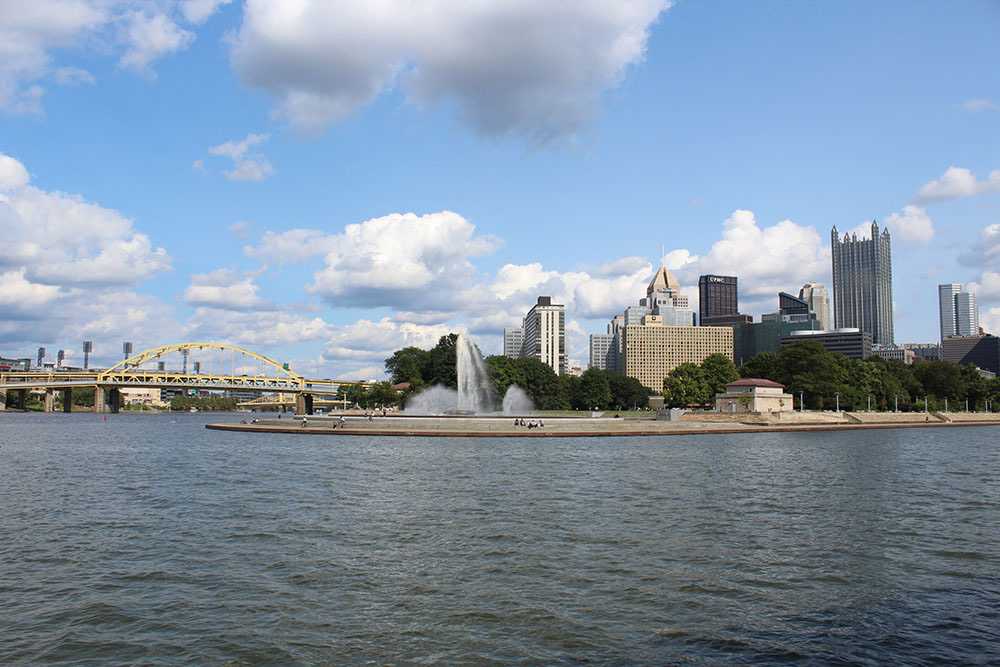 Photo by Frankie Bonamo, 2021. Features the Pittsburgh skyline featuring Point State Park Fountain in the center, with a yellow bridge to the left and several city skyscrapers to the right 